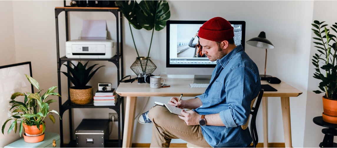 Guy sitting at desk with computer writing in notebook